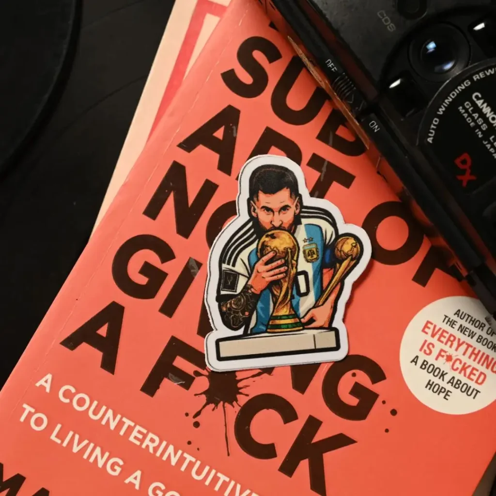 A sticker of Lionel Messi celebrating his World Cup victory, resting on an orange book cover next to a vintage camera.