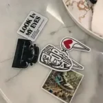A group of graphic and text-based stickers, including one that says "LOOK IN MY EYES," resting on a marble surface next to a white decorative tray.